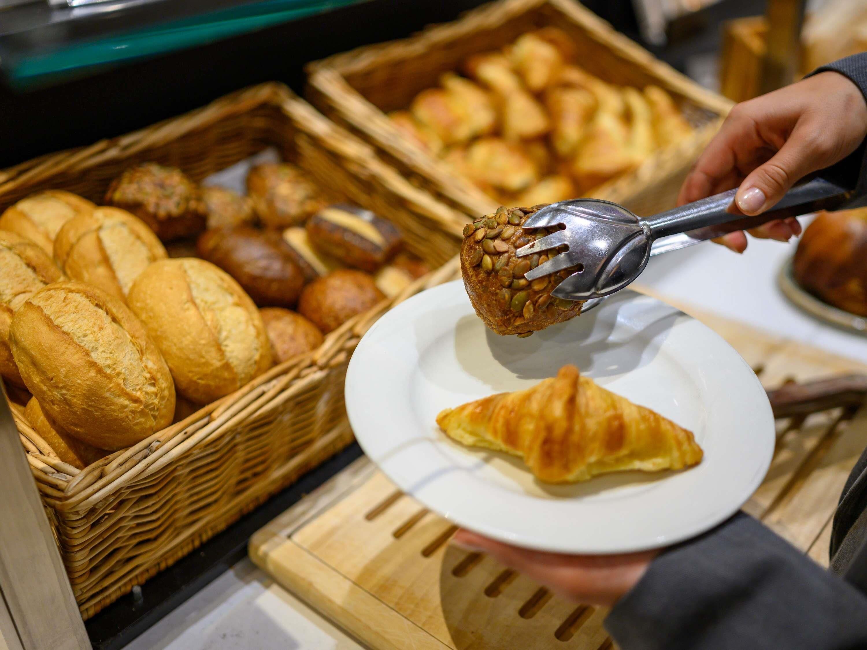 A hand holding a spoon with a bread roll, a plate with a croissant, and baskets of various baked goods.