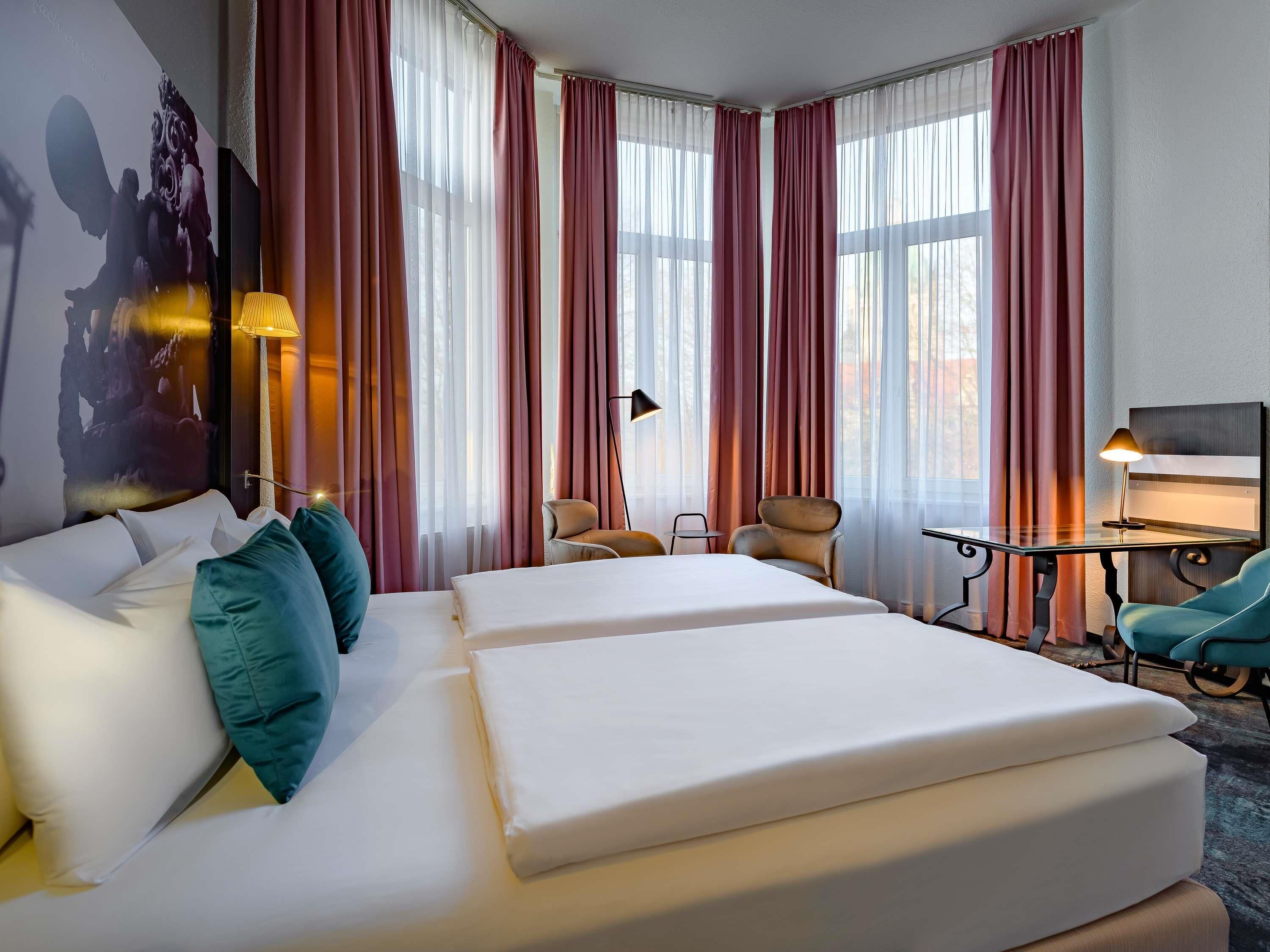 A hotel room with two beds, a desk, chairs, and large windows with curtains.