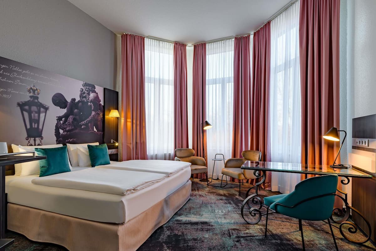 Une chambre d’hôtel avec un grand lit, deux chaises, une petite table, un bureau et une grande fenêtre avec des rideaux.