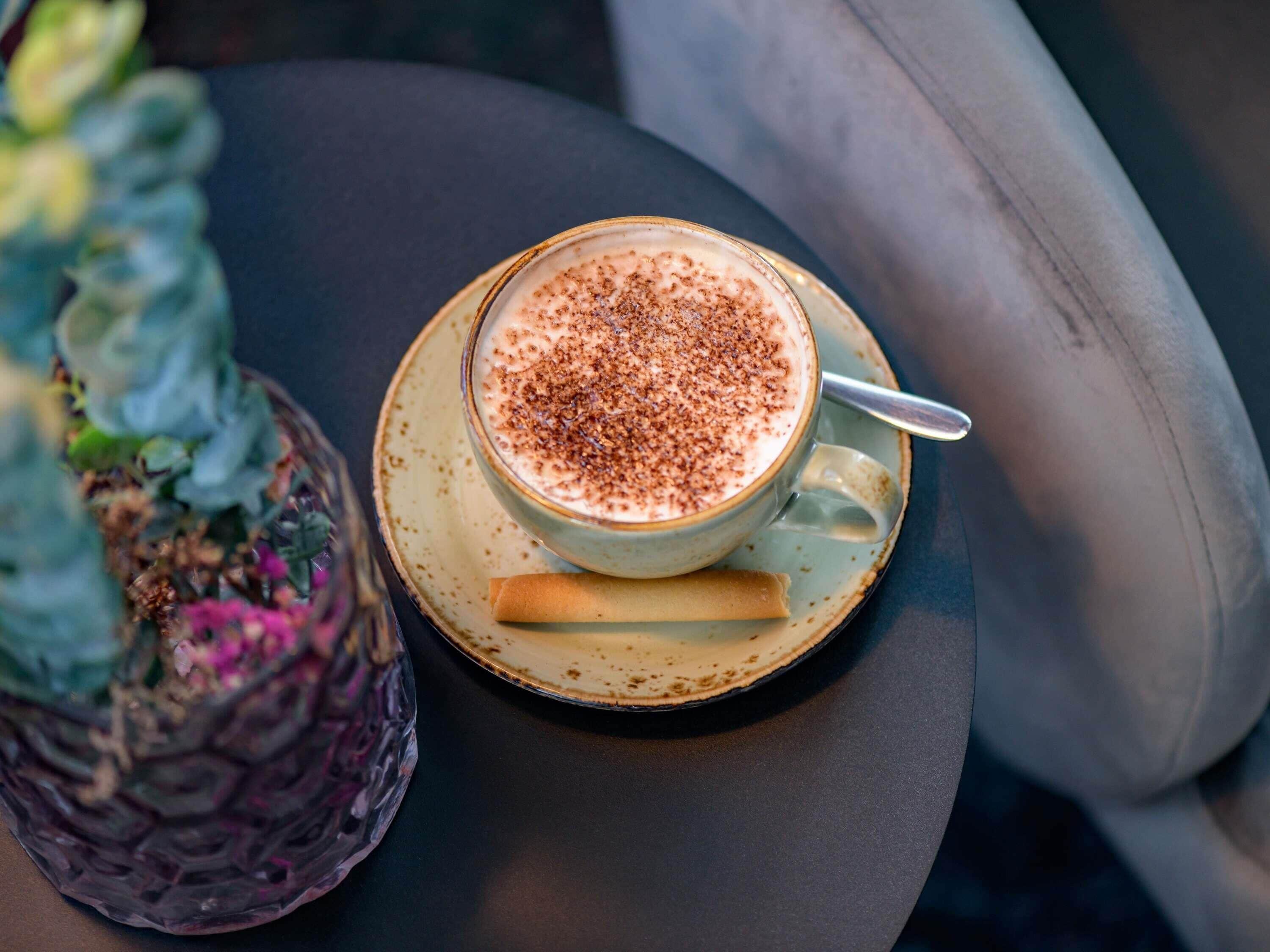 A cup of coffee with a sprinkle of cocoa on a saucer, placed on a table with a cushion.