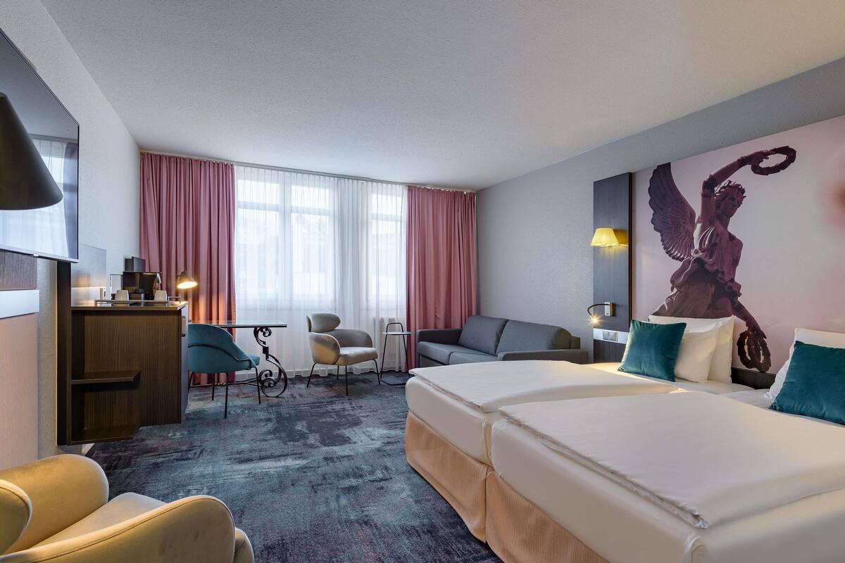 Een hotelkamer met een groot bed, een bureau met een computer, een stoel, een bank en een groot raam met gordijnen.