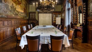 A dining room with a long table, chairs, a chandelier, and a large painting on the wall.