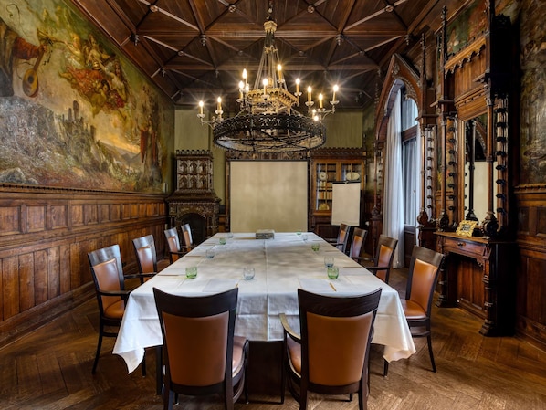 A dining room with a long table, chairs, a chandelier, and a large painting on the wall.