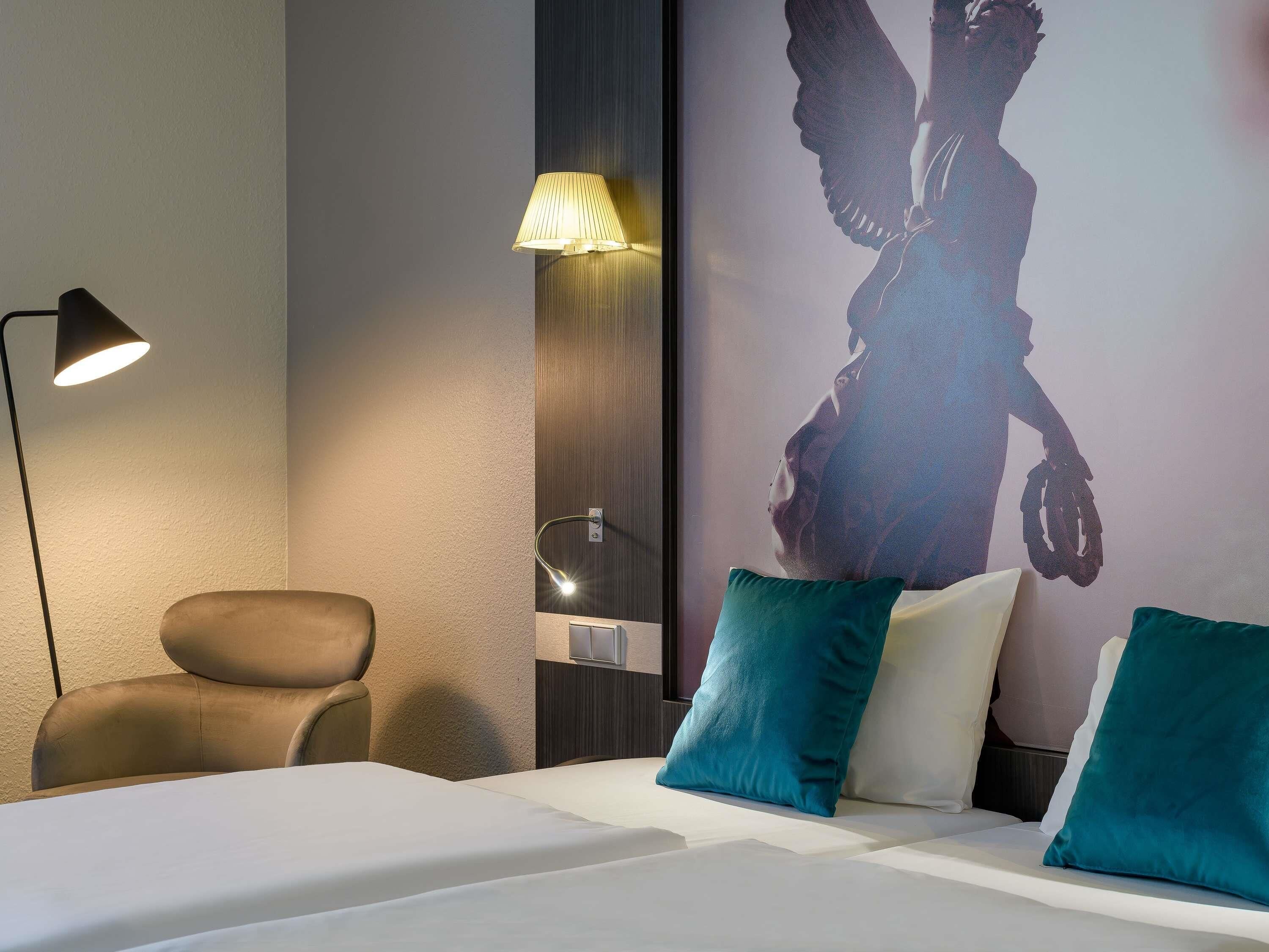 A hotel room with a bed, a desk, a chair, a lamp, and a wall with a silhouette artwork.