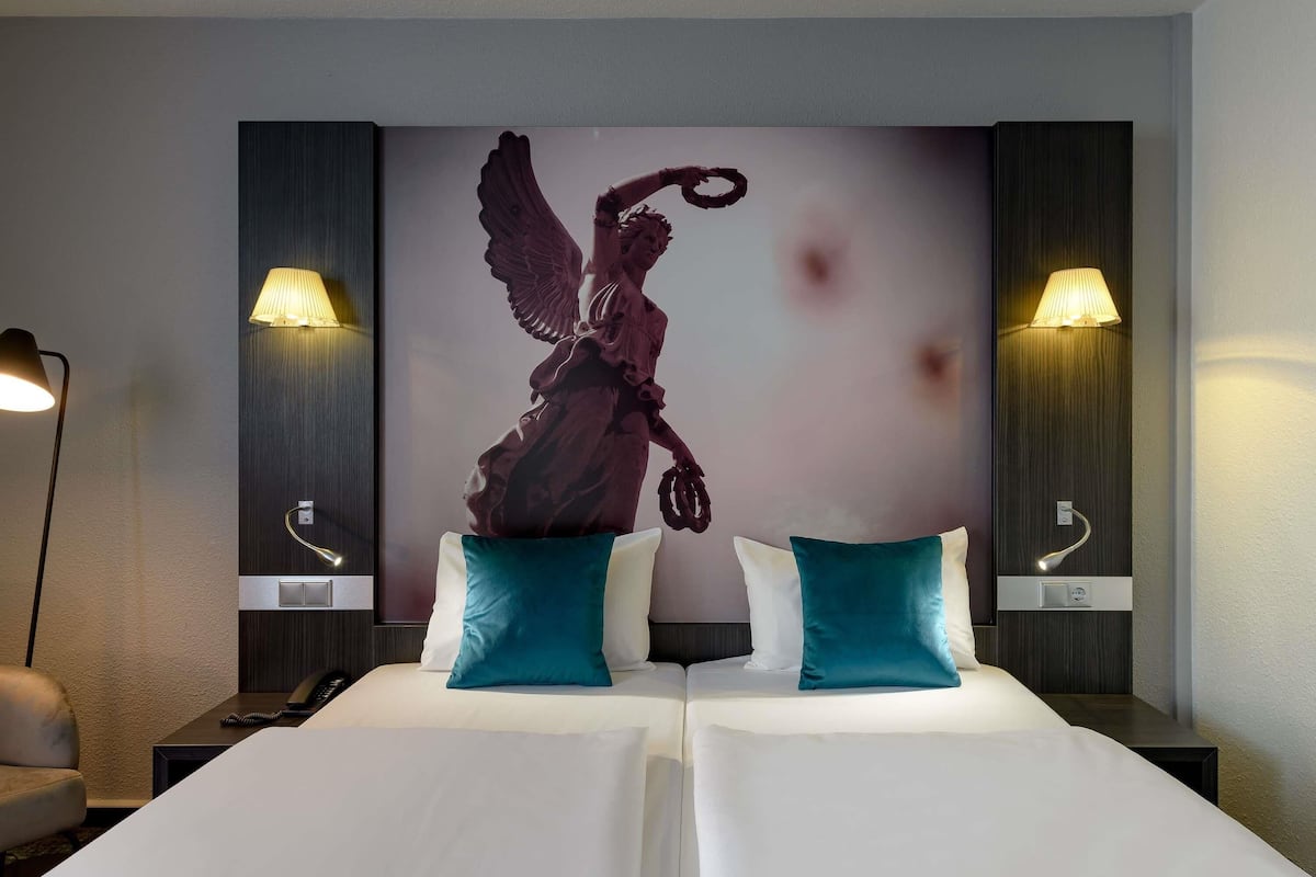 Hotelkamer met een groot bed, twee kussens, een schilderij van een engel en twee bedlampjes.
