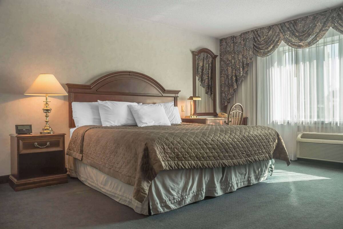 A neatly made bed with a headboard, a bedside table with a lamp, a mirror, and a window with curtains.