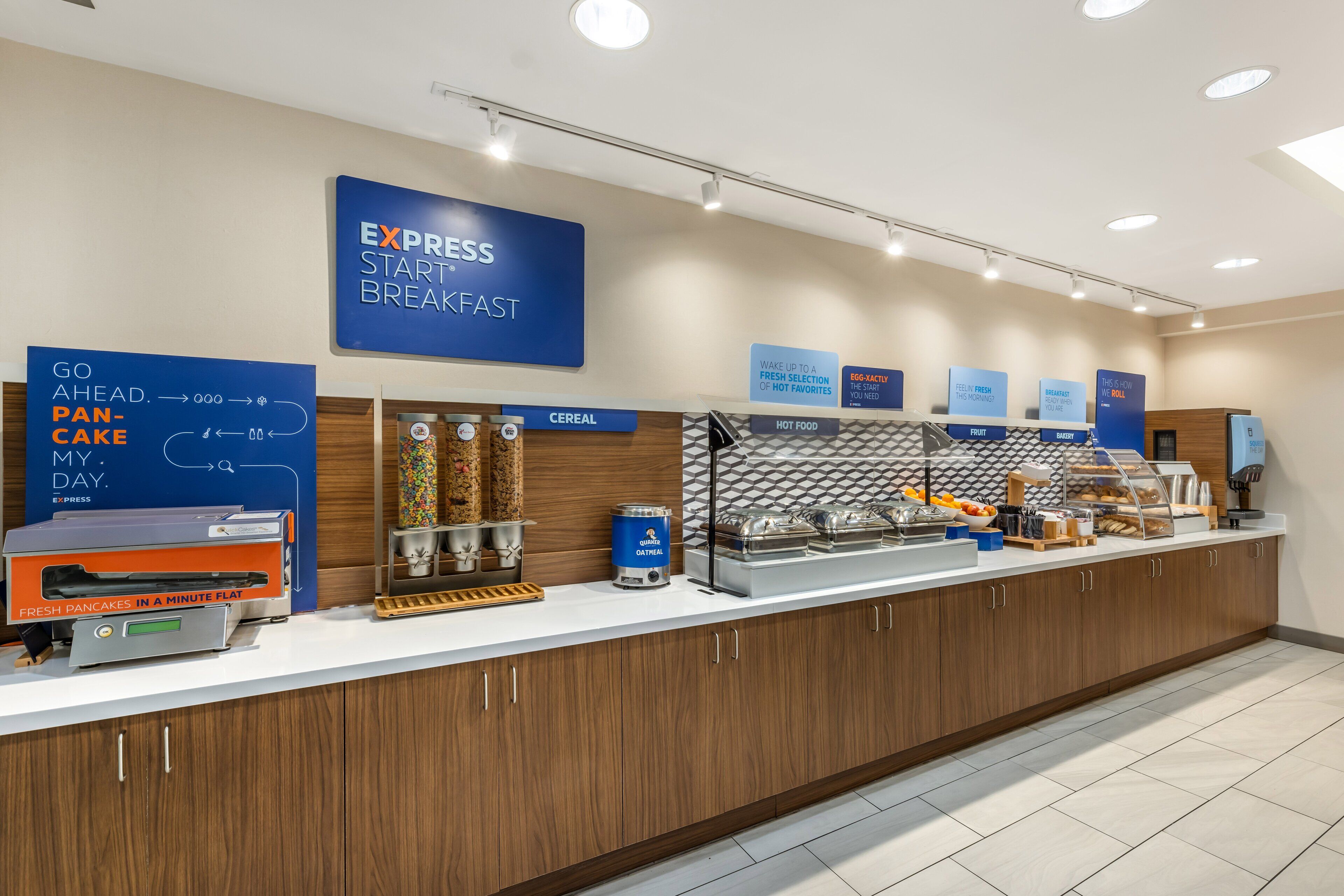 Un área de desayuno con letreros que indican 'EXPRESS START BREAKFAST' y 'GO AHEAD. PANCAKE MY DAY'. Hay secciones para cereales, comida caliente y fruta.