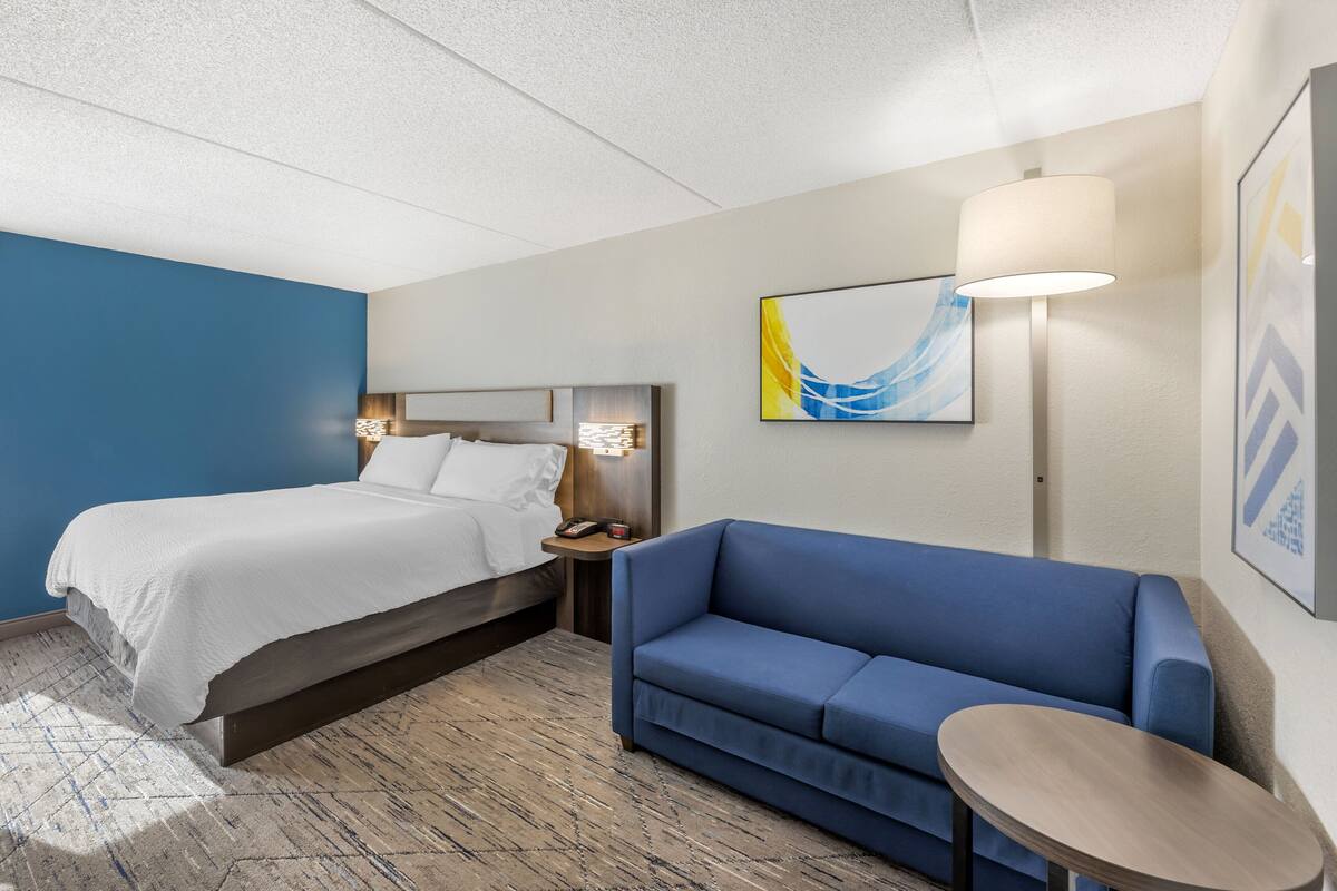 A hotel room with a bed, a blue sofa, a small table, a nightstand, and a lamp.