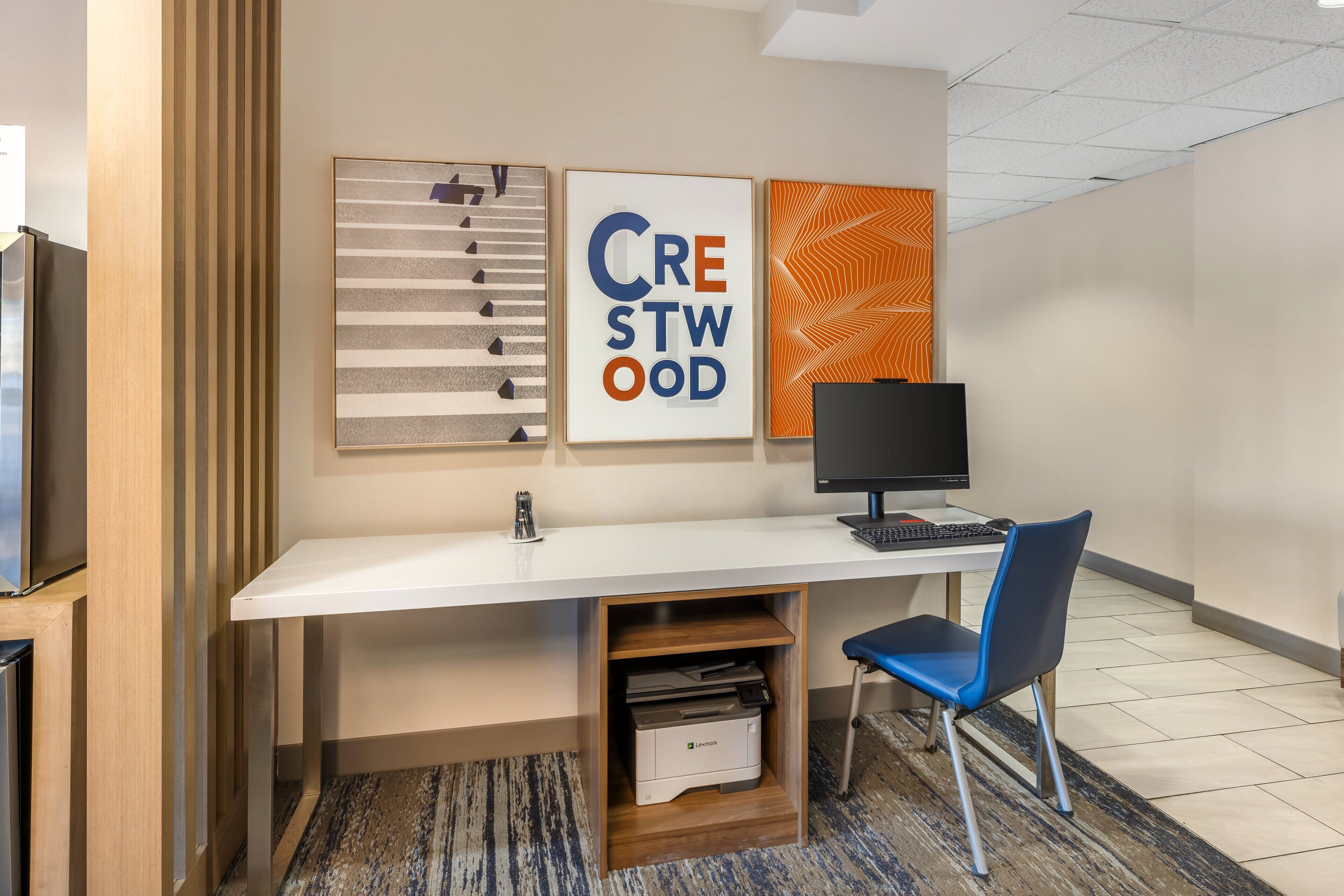 A modern office space with a white desk, a blue chair, a computer monitor, and framed artwork on the wall.