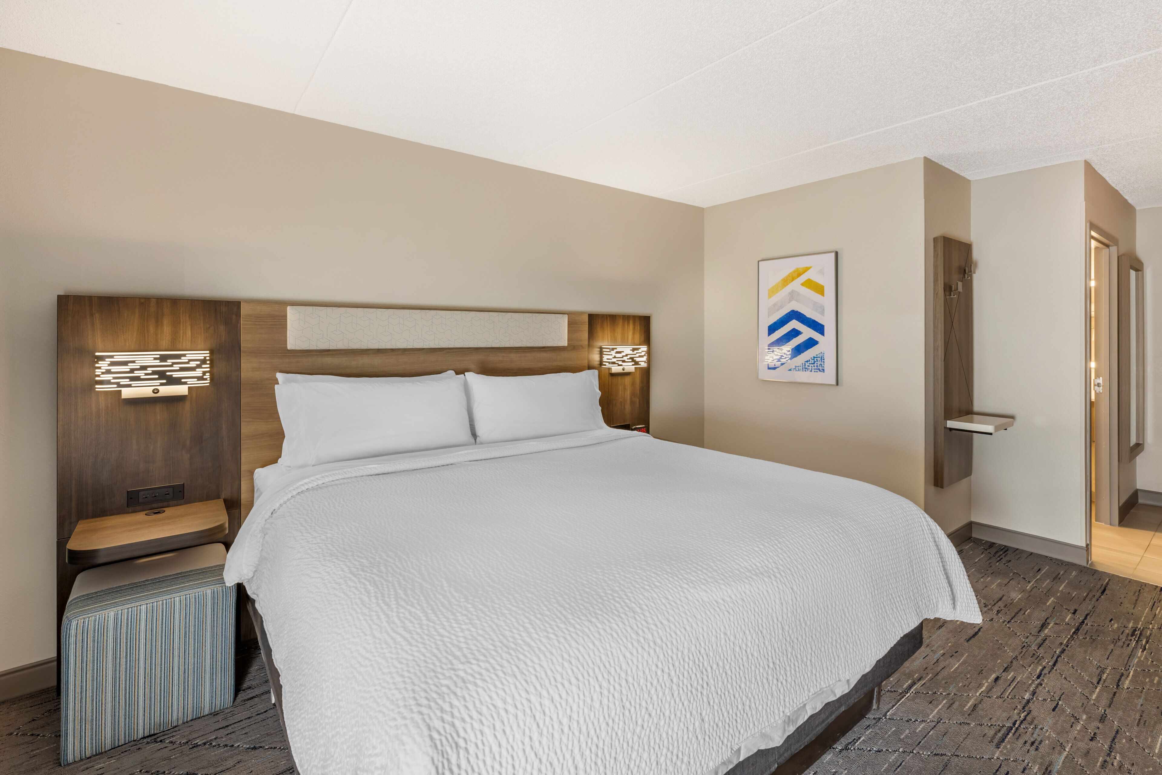 A hotel room with a large bed, a nightstand, a wall-mounted shelf, and a door to the bathroom.