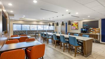 Un área de comedor moderna con varias mesas y sillas, una estación de café y un ventanal con vistas al exterior.
