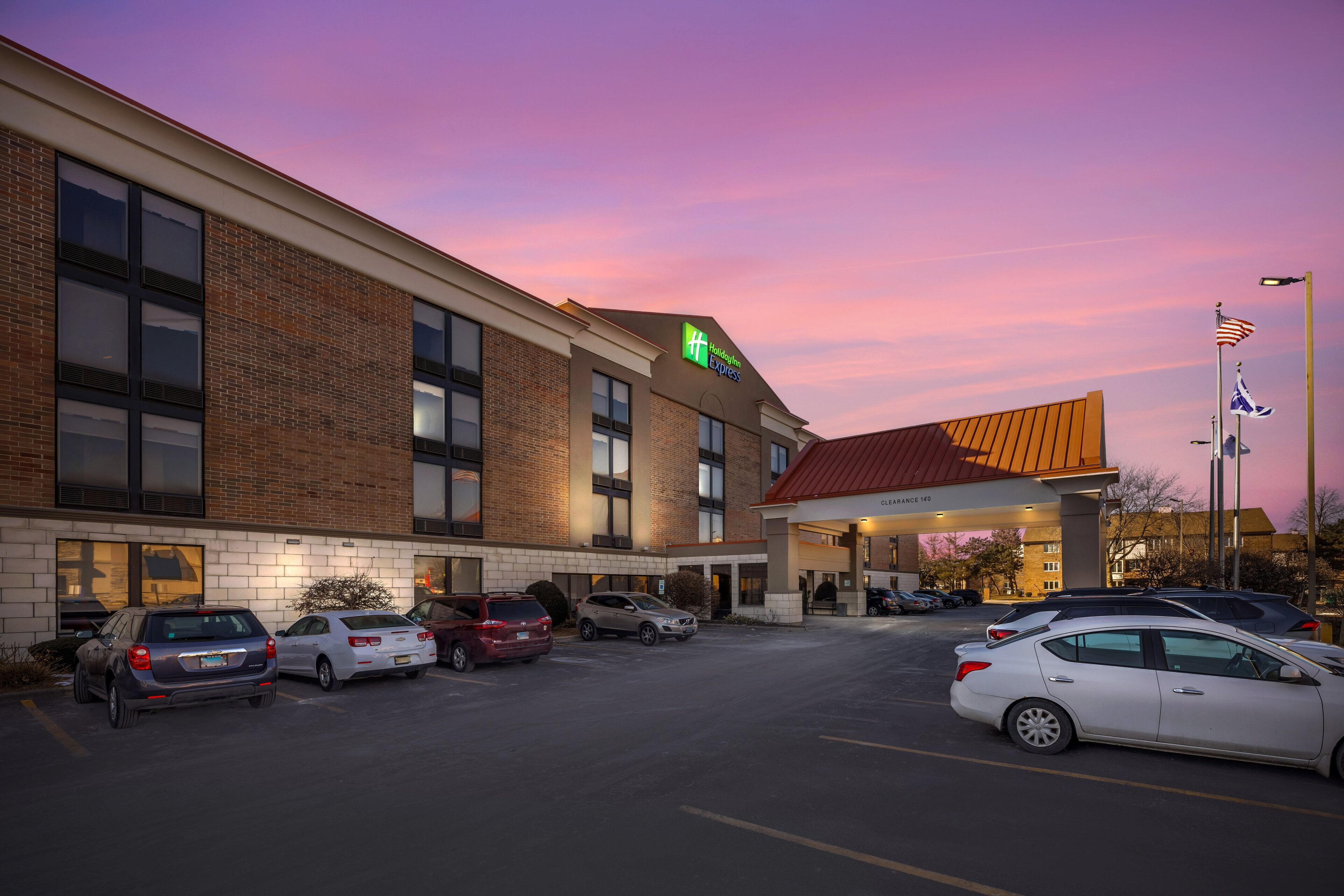 A hotel with a brick facade, a red roof, and a parking lot with several cars.
