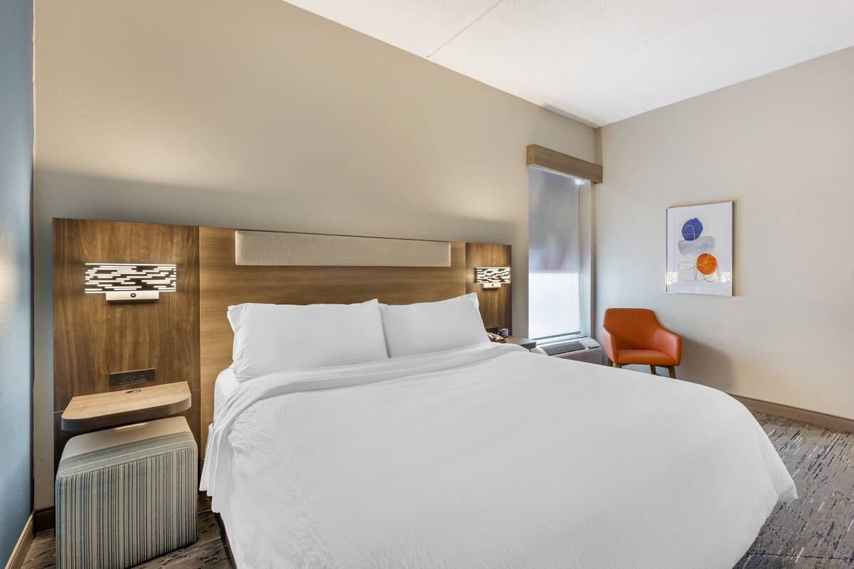 A modern hotel room with a large bed, a wooden headboard, a small side table, a chair, and a large window.