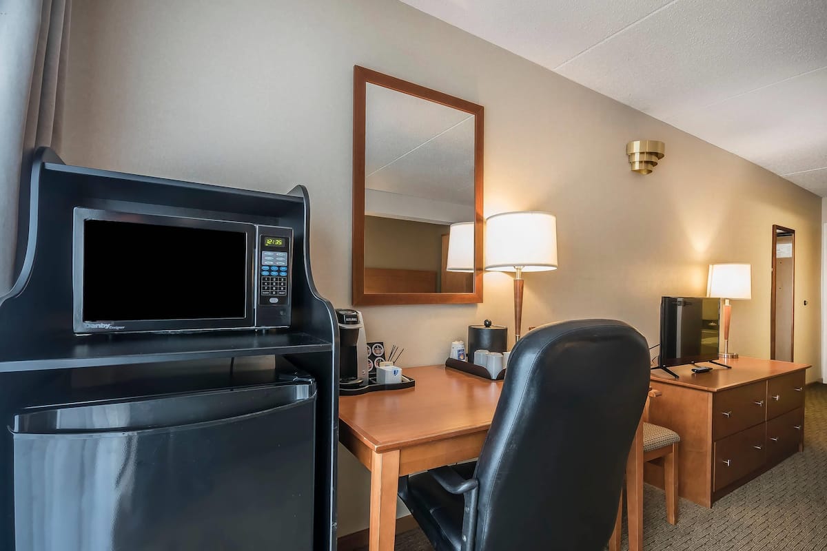 A hotel room with a desk, a chair, a microwave, a coffee maker, a lamp, a TV, and a mirror.