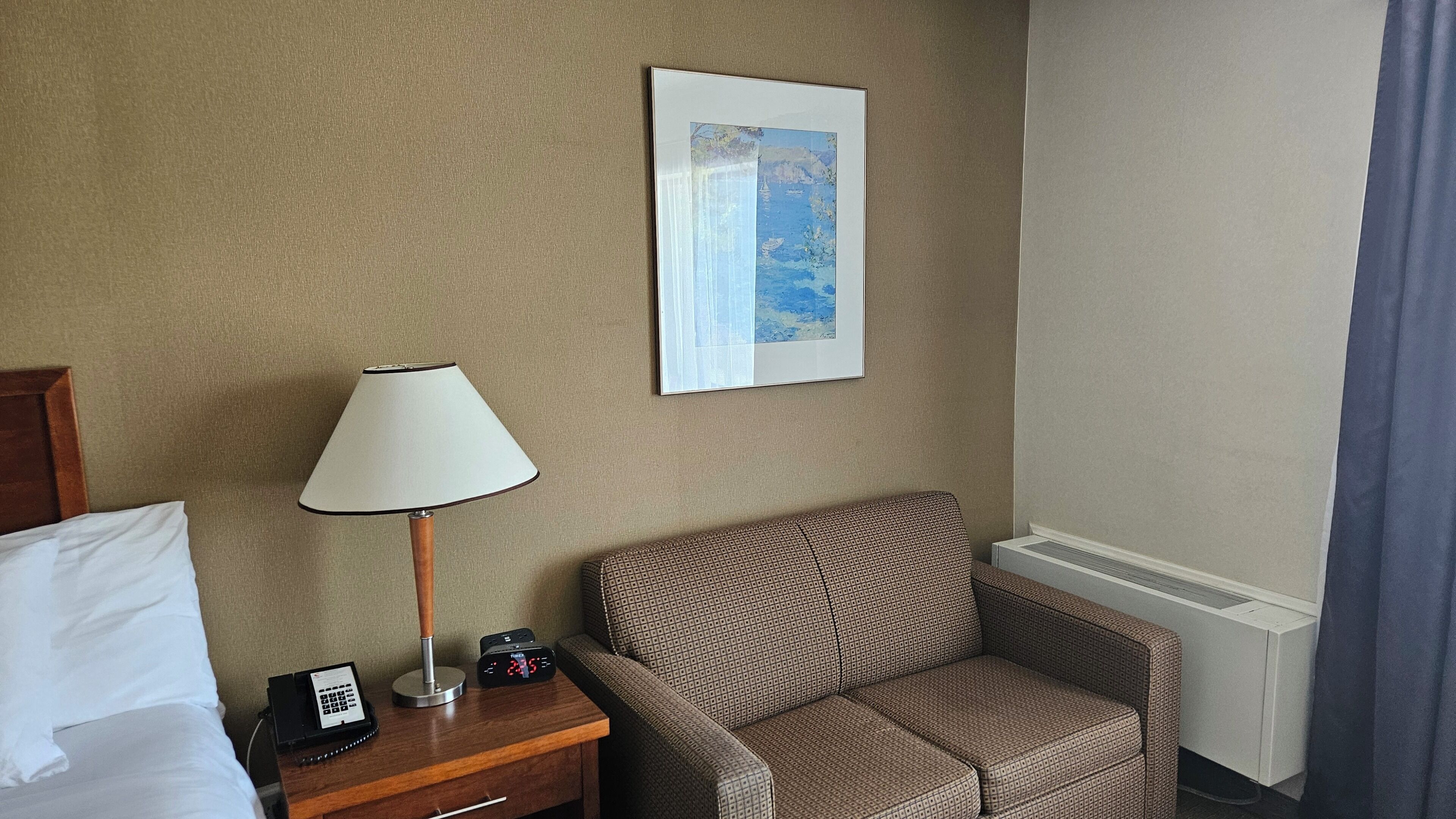 Habitación de hotel con cama, mesita de noche de madera con lámpara, reloj digital, teléfono y sillón estampado.
