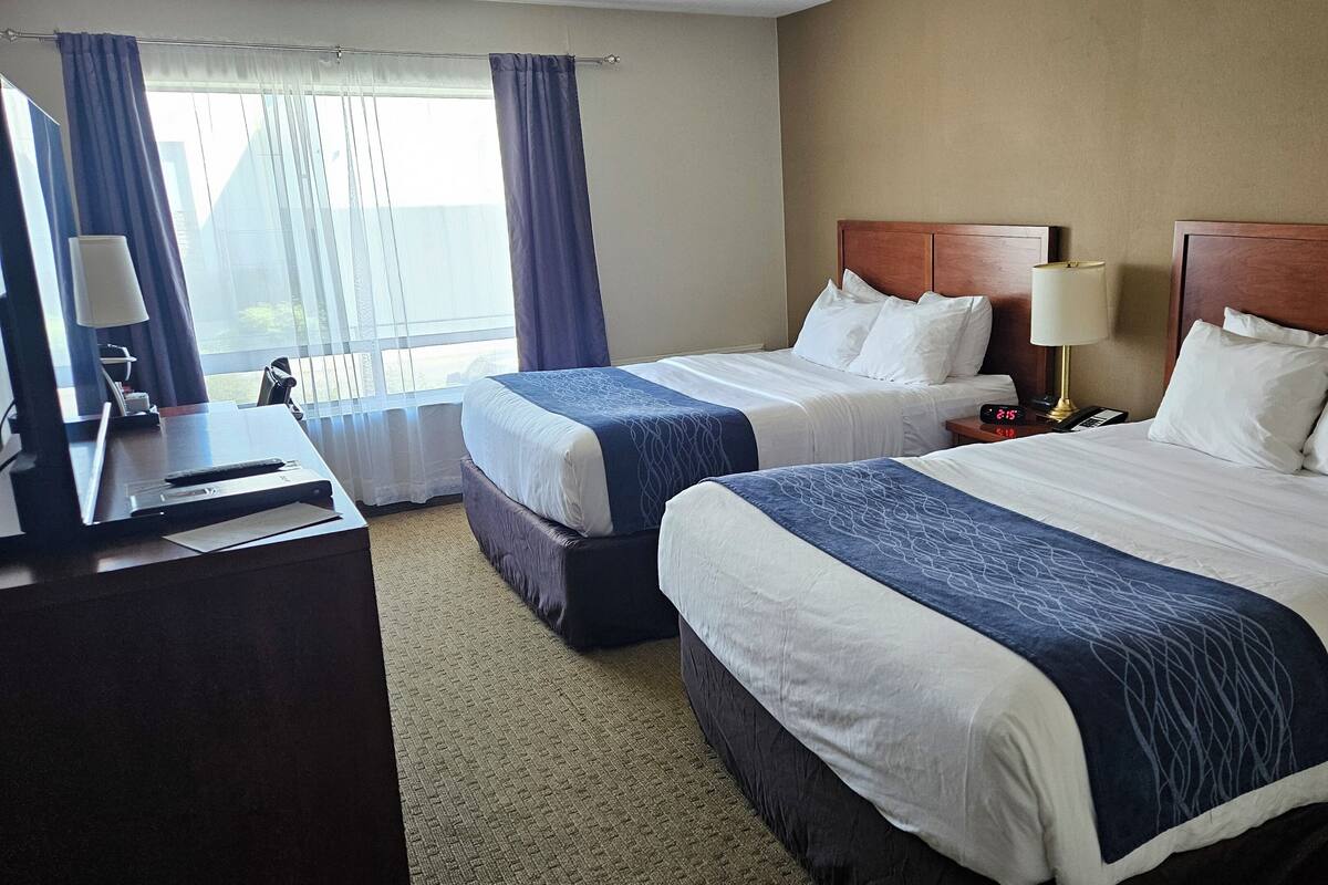 A hotel room with two beds, a desk with a lamp, a chair, and a window with curtains.