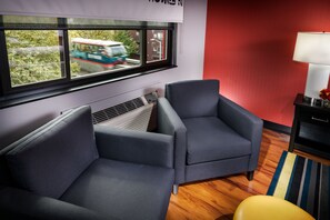 A modern living room with two grey armchairs, a window with a view of a moving train, a red accent wall, and a lamp on a side table.