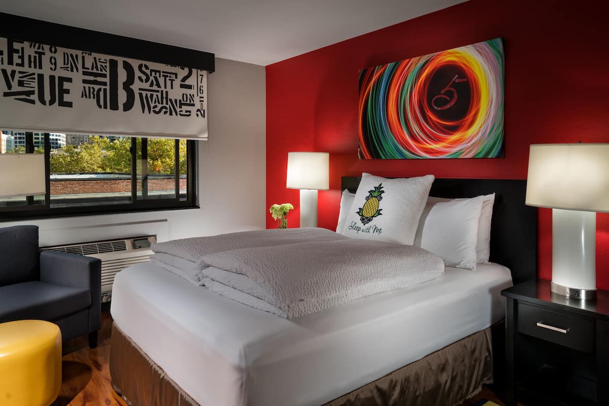 A modern hotel room with a large bed, two bedside lamps, a colorful abstract painting, and a window with a view of trees.