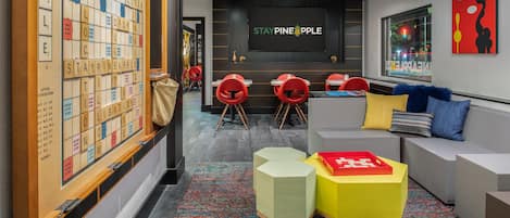 A modern lounge area with a Scrabble board, a large screen displaying "STAY PINEAPPLE", and a seating area with colorful chairs and sofas.