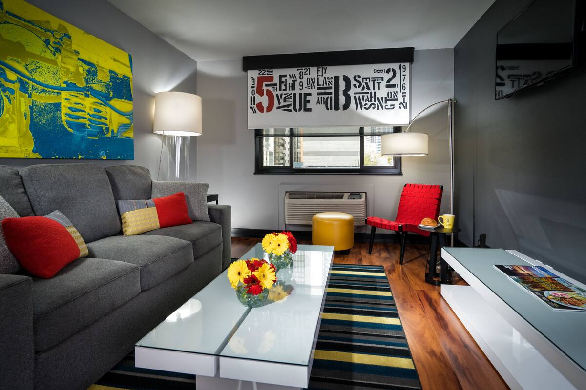 A modern living room with a grey sofa, red and yellow cushions, a glass coffee table with flowers, a red chair, and a large abstract painting on the wall.
