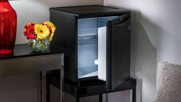 A compact living space with a modern black refrigerator on a stand, a glass vase with flowers on a table, and a grey sofa with a red pillow.