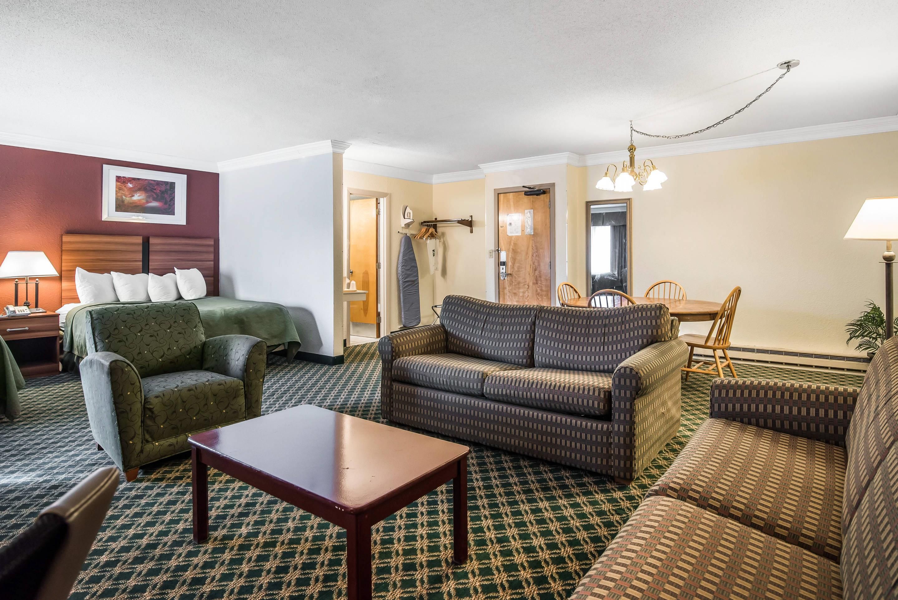 A hotel room with a green patterned sofa, a wooden coffee table, a patterned armchair, and a dining area with a wooden table and chairs.