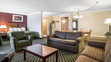 A hotel room with a green patterned sofa, a wooden coffee table, a patterned armchair, and a dining area with a wooden table and chairs.