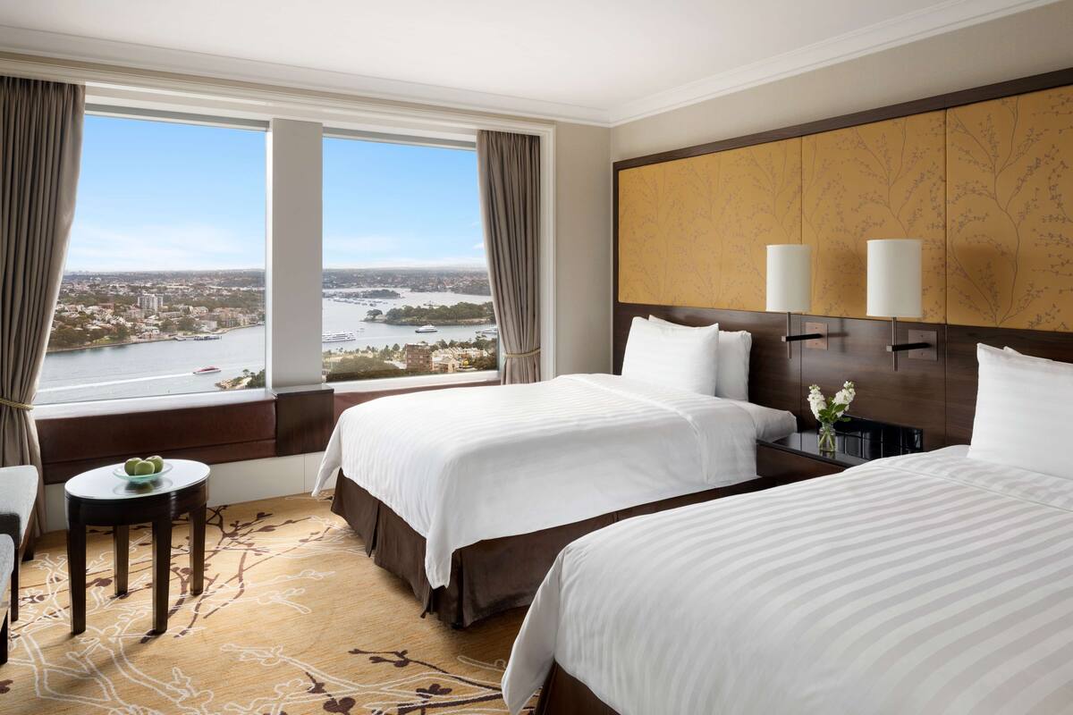 Ein Hotelzimmer mit zwei Betten, Blick auf Stadt und Fluss, einem kleinen Tisch mit Blumenvase und einem großen Holzkopfteil mit zwei Wandleuchten.