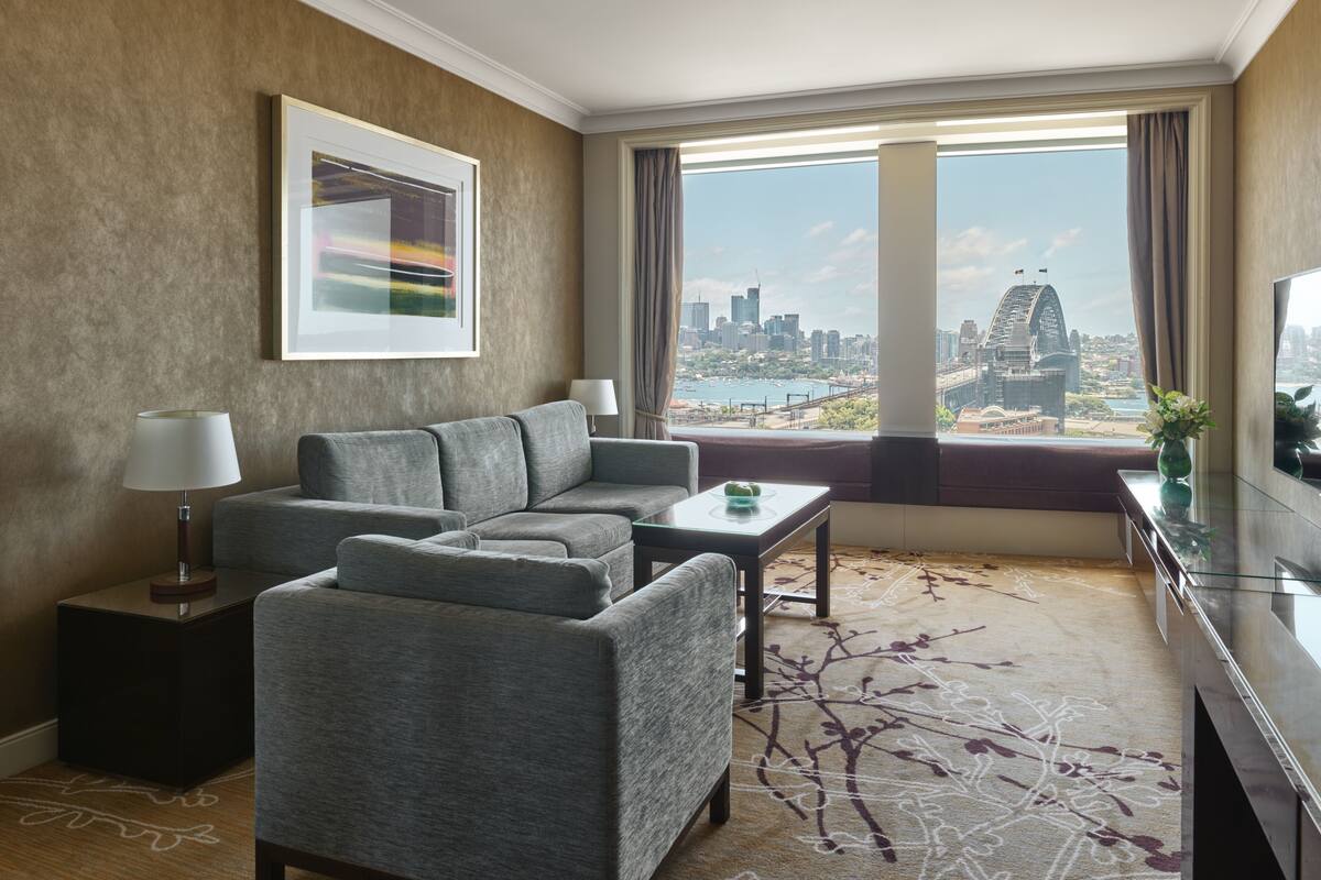A modern hotel room with a grey sofa set, a small table, a lamp, a TV, and a large window offering a city view.