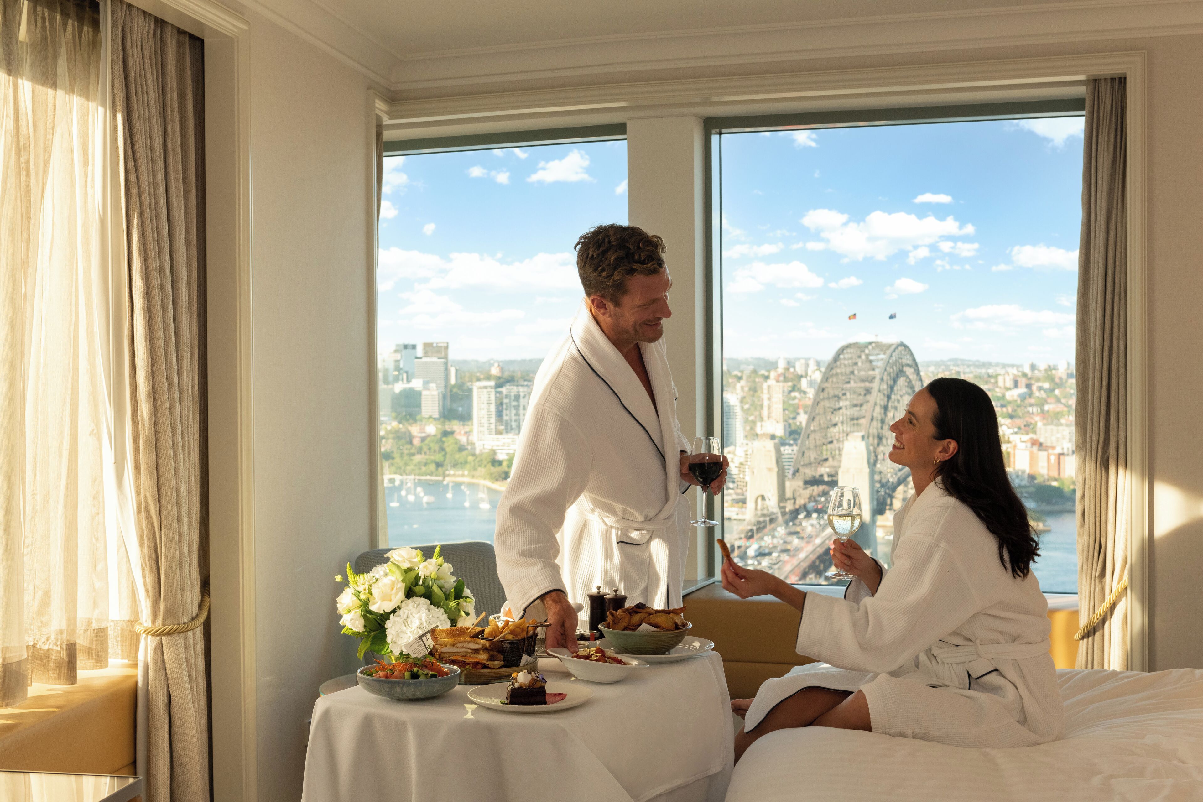 Una pareja en una habitación de hotel con una mesa lista para el desayuno, vistas a la ciudad y un gran ventanal.