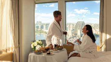 Una pareja en una habitación de hotel con una mesa lista para el desayuno, vistas a la ciudad y un gran ventanal.