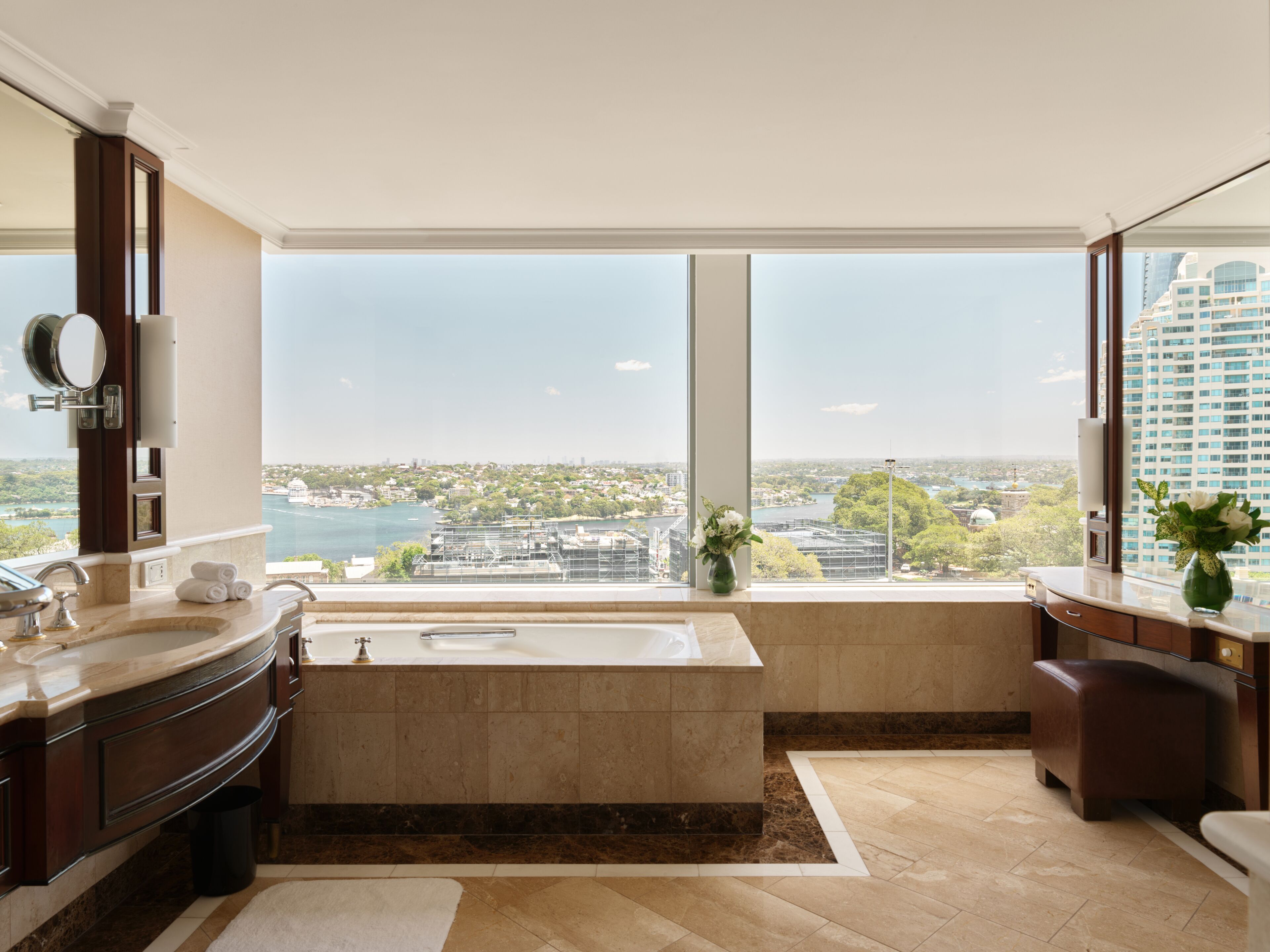 Ein modernes Badezimmer mit einer großen Badewanne, einem Waschtisch inklusive Spiegel und Blick auf eine Stadtlandschaft durch große Fenster.
