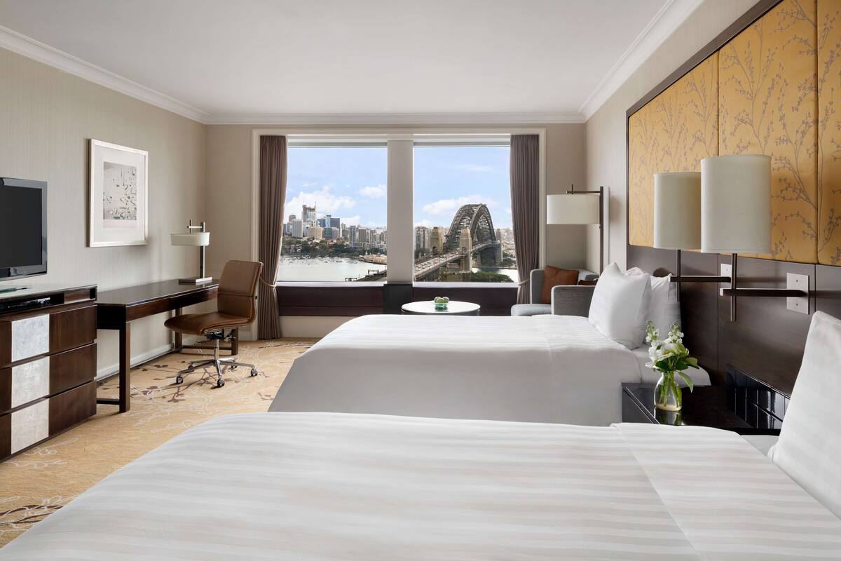 Ein Hotelzimmer mit zwei Betten, einem Schreibtisch, einem Sessel, einem Fernseher und Blick auf Stadt und Brücke.