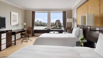 A hotel room with two beds, a desk, a chair, a TV, and a view of a cityscape and bridge.