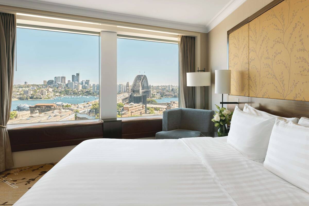 Ein Hotelzimmer mit einem großen Bett, Blick auf eine Stadtlandschaft und eine Brücke, einem Sessel und einer Lampe.