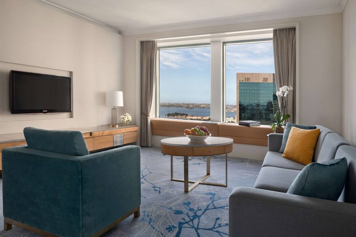 A modern hotel room with a sofa, a round table with a fruit bowl, a TV mounted on the wall, and a view of the city and water through large windows.