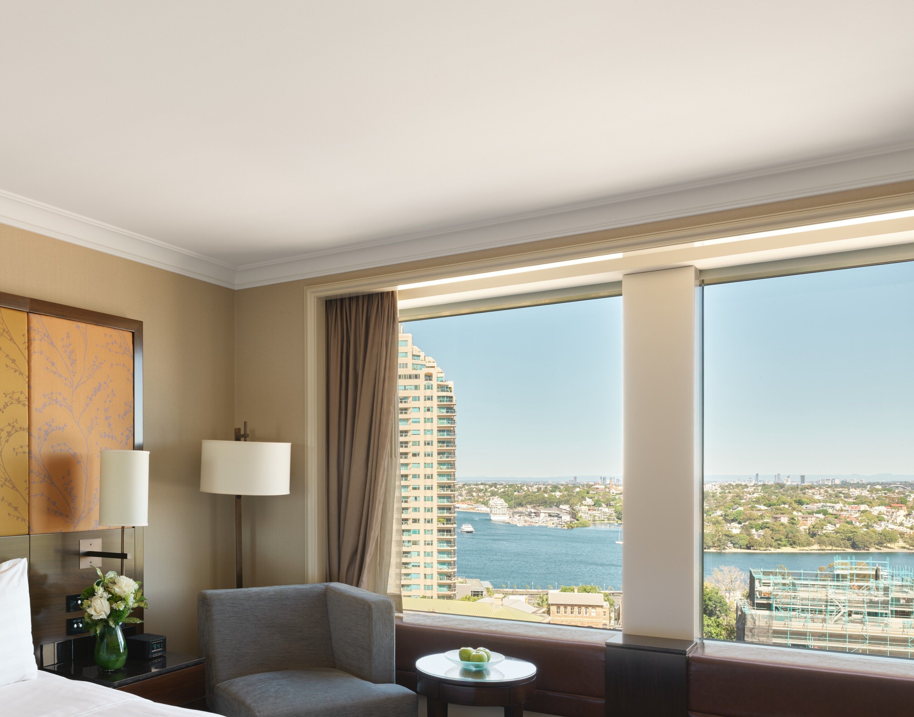A hotel room with a large window, a bed, a chair, a small table, and a lamp.