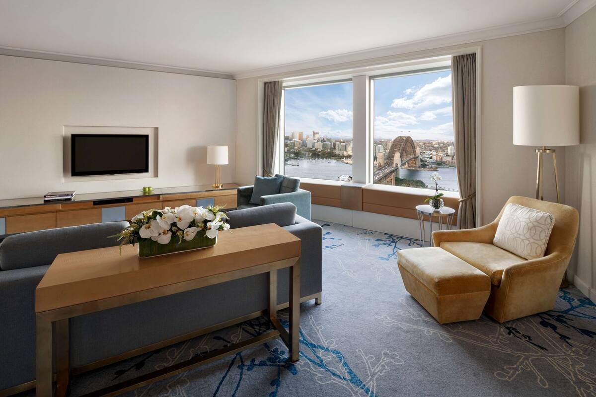 A modern hotel room with a sofa, armchair, and a dining table with a vase of flowers. A TV is mounted on the wall, and there is a view of a cityscape through large windows.