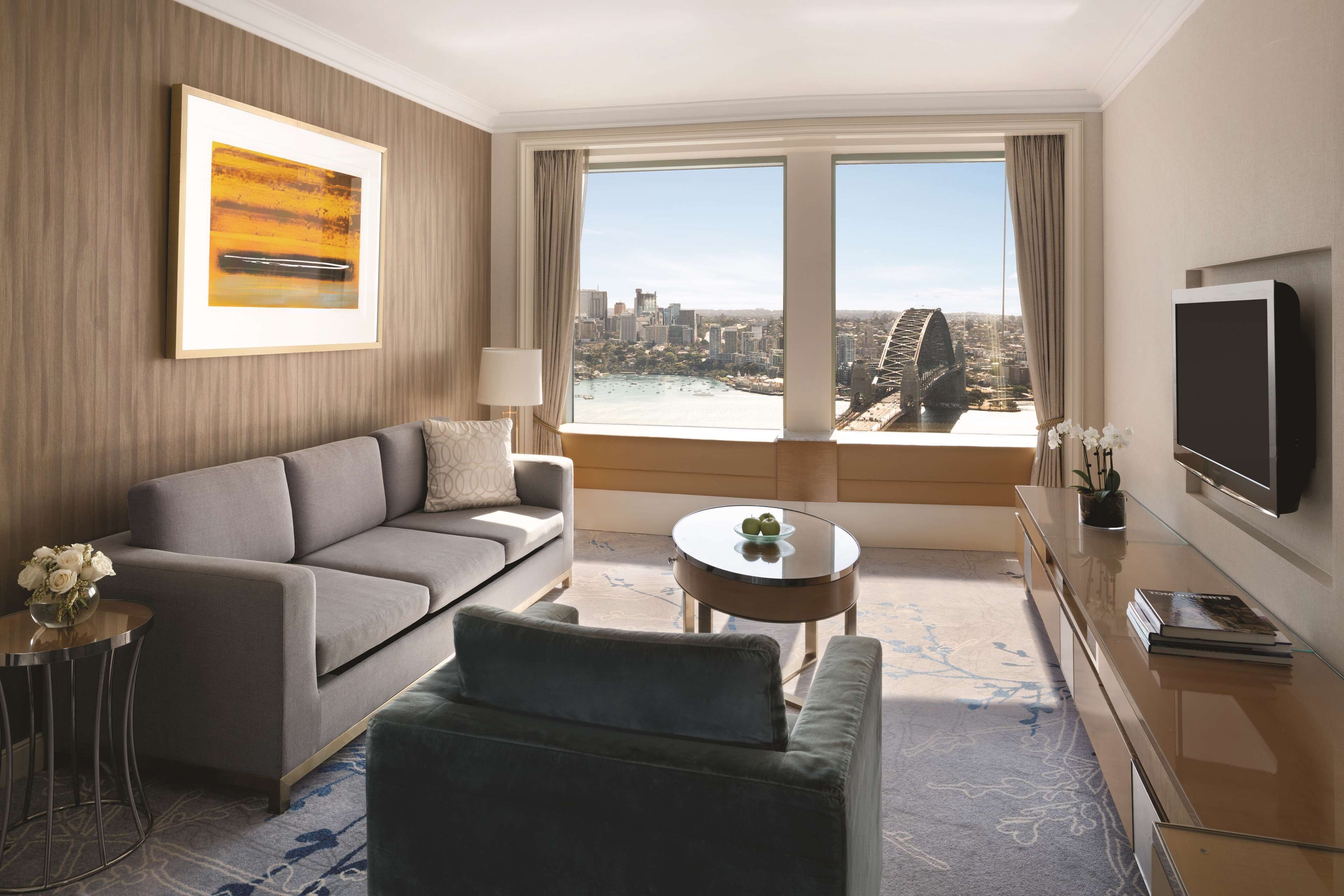 A modern hotel room with a sofa, a coffee table, a TV, and a large window with a city view.