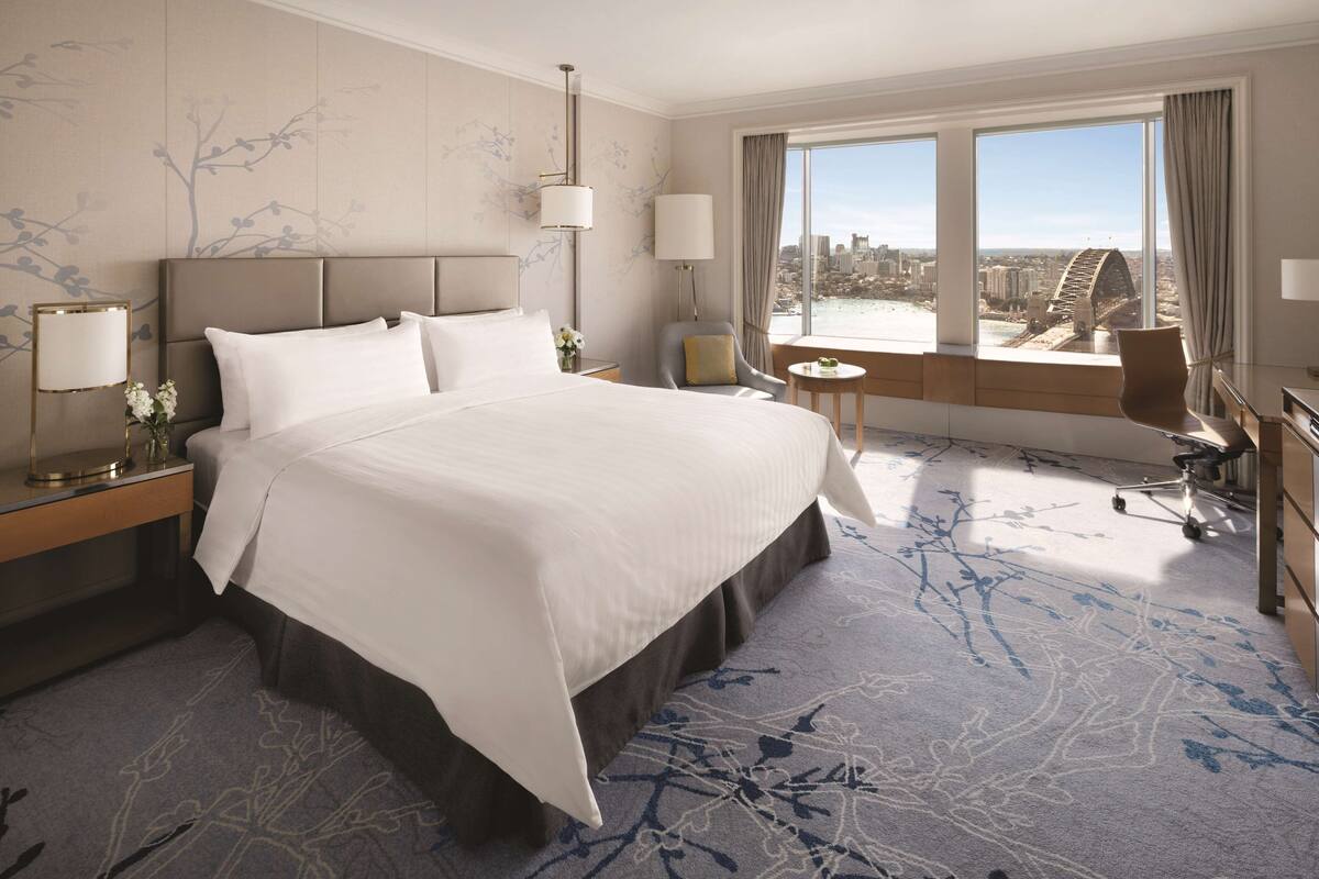 A hotel room with a large bed, a desk, a chair, and a view of a cityscape and bridge.