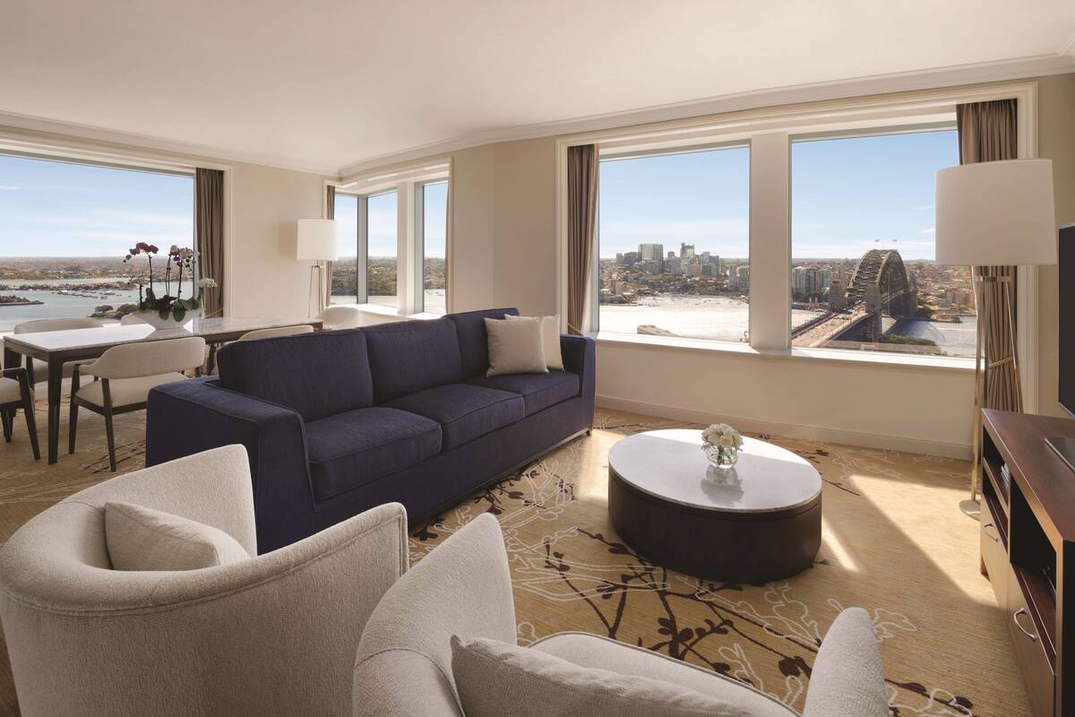 A modern hotel room with a large sofa, a dining table, and a view of a cityscape and bridge.