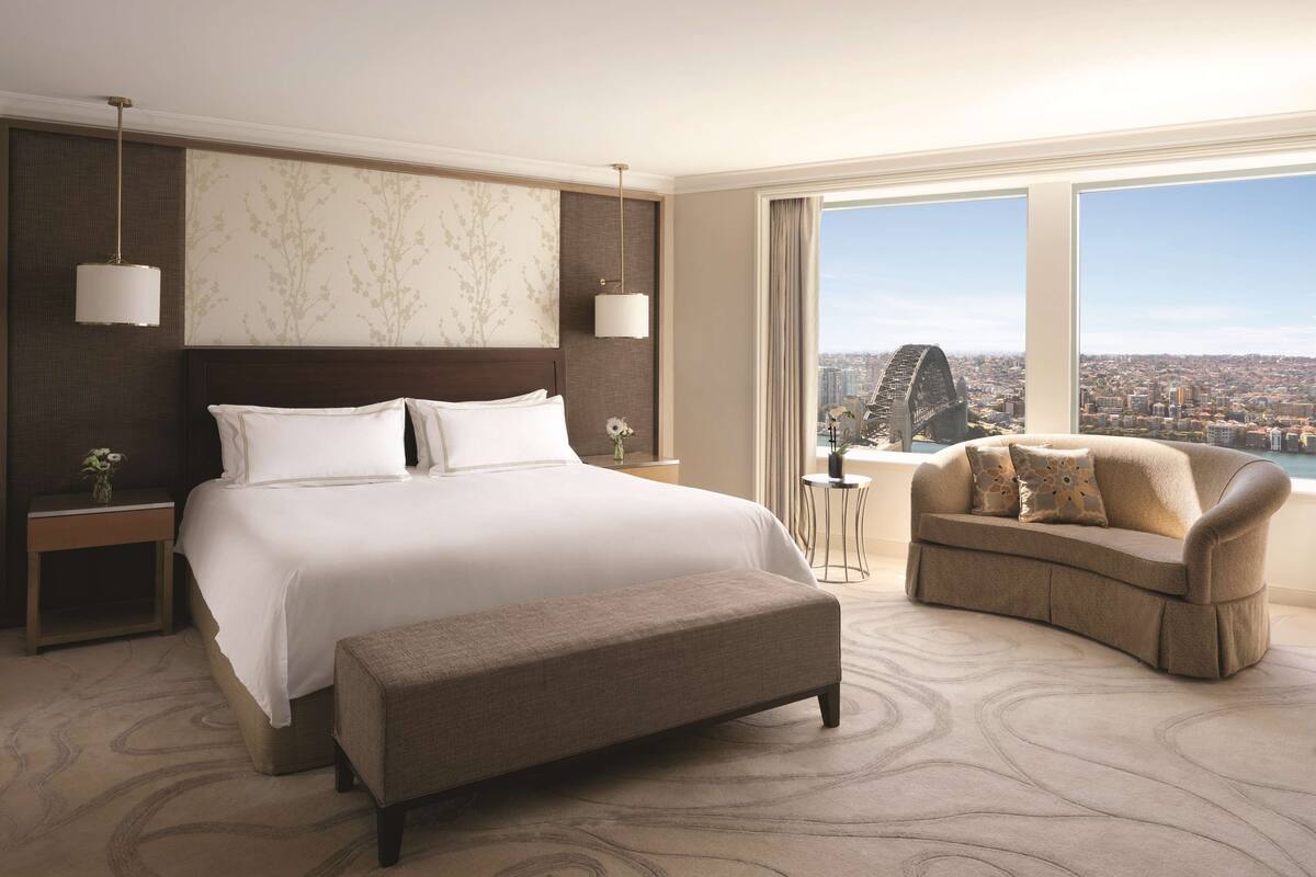 A hotel room with a large bed, a sofa, a bench, and a view of a cityscape and a bridge.