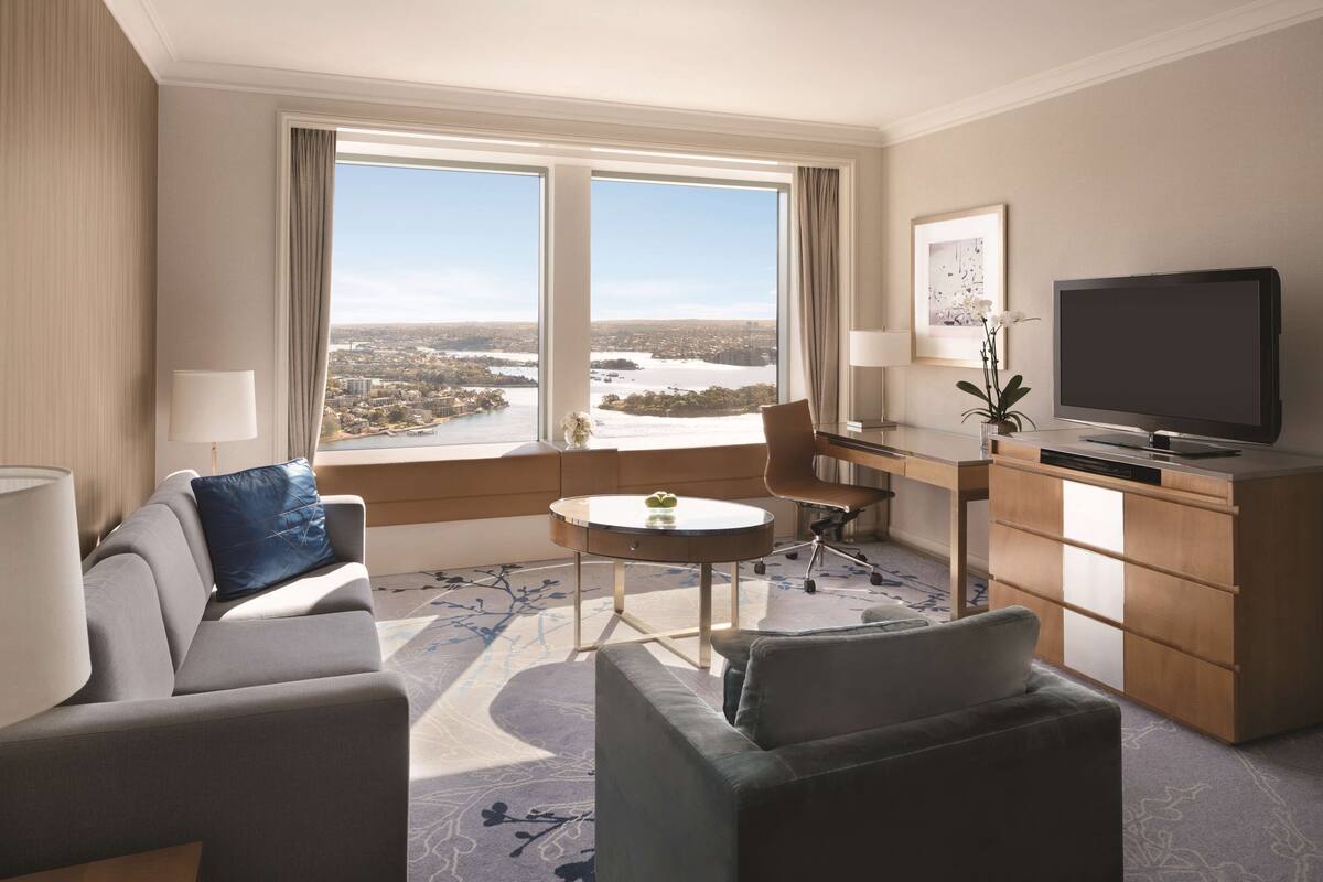 A hotel room with a large window offering a city view, a desk with a chair, a television, and a sofa.