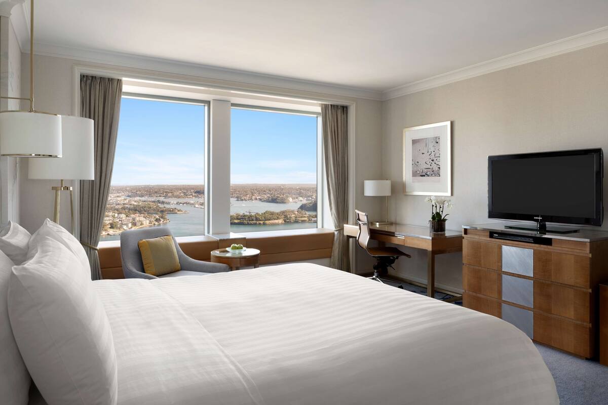 A hotel room with a large bed, a desk with a TV, a chair, a small table with a plant, and a view of a cityscape through large windows.