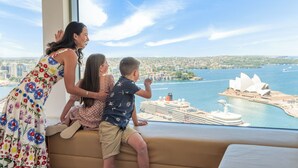 A family of three on a balcony overlooking a cityscape with a large cruise ship and the Sydney Opera House. - Shangri-La Sydney (The Rocks)