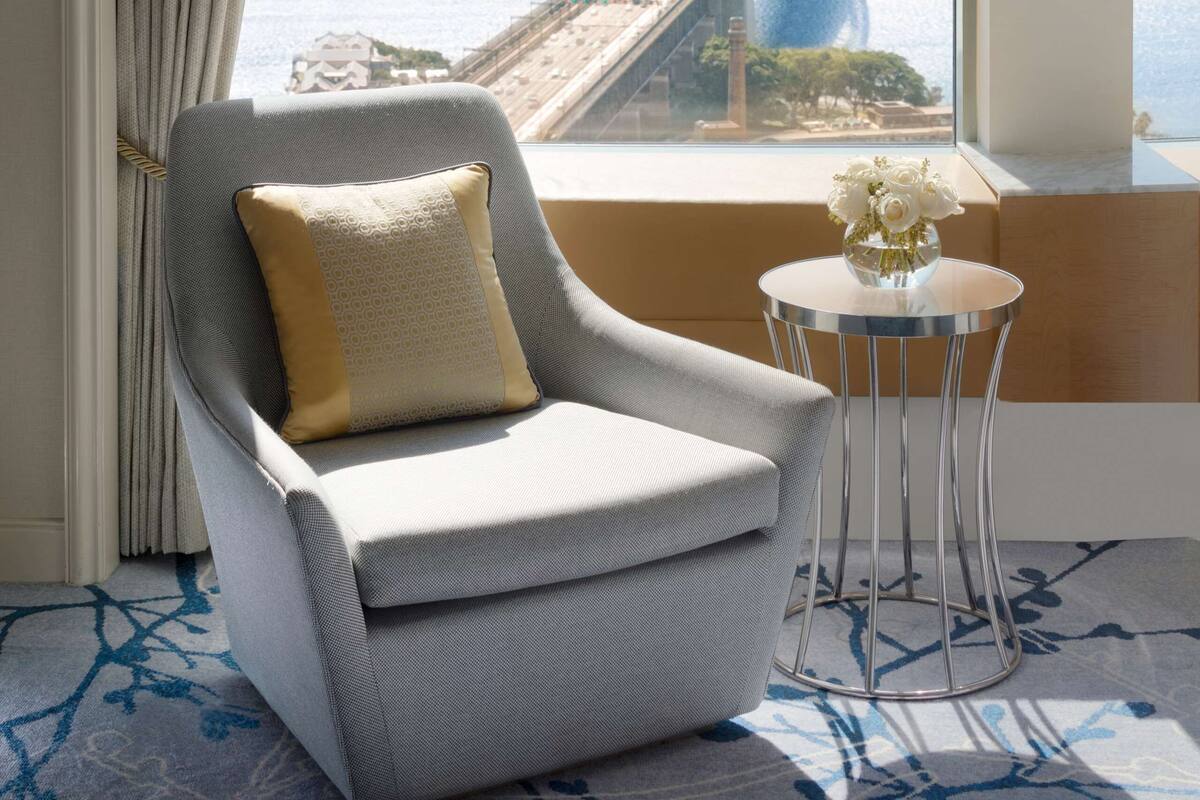 A room with a view of a bridge and cityscape, a grey armchair with a cushion, and a small table with a vase.