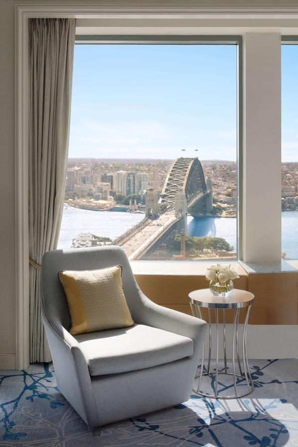 A room with a view of a bridge and cityscape, a grey armchair with a cushion, and a small table with a vase.