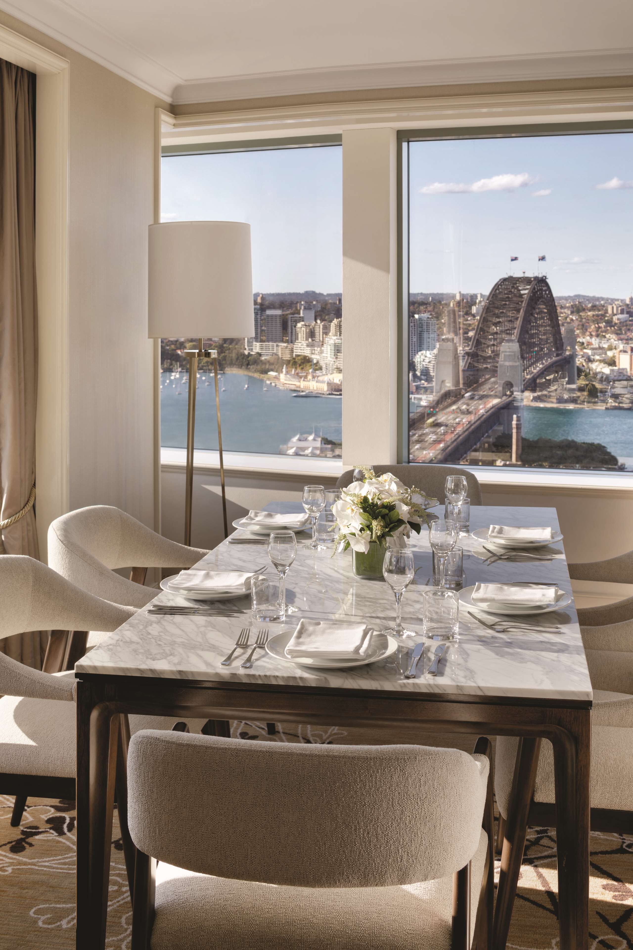 A dining area with a table set for four, a vase of flowers, and a view of a cityscape with a bridge.
