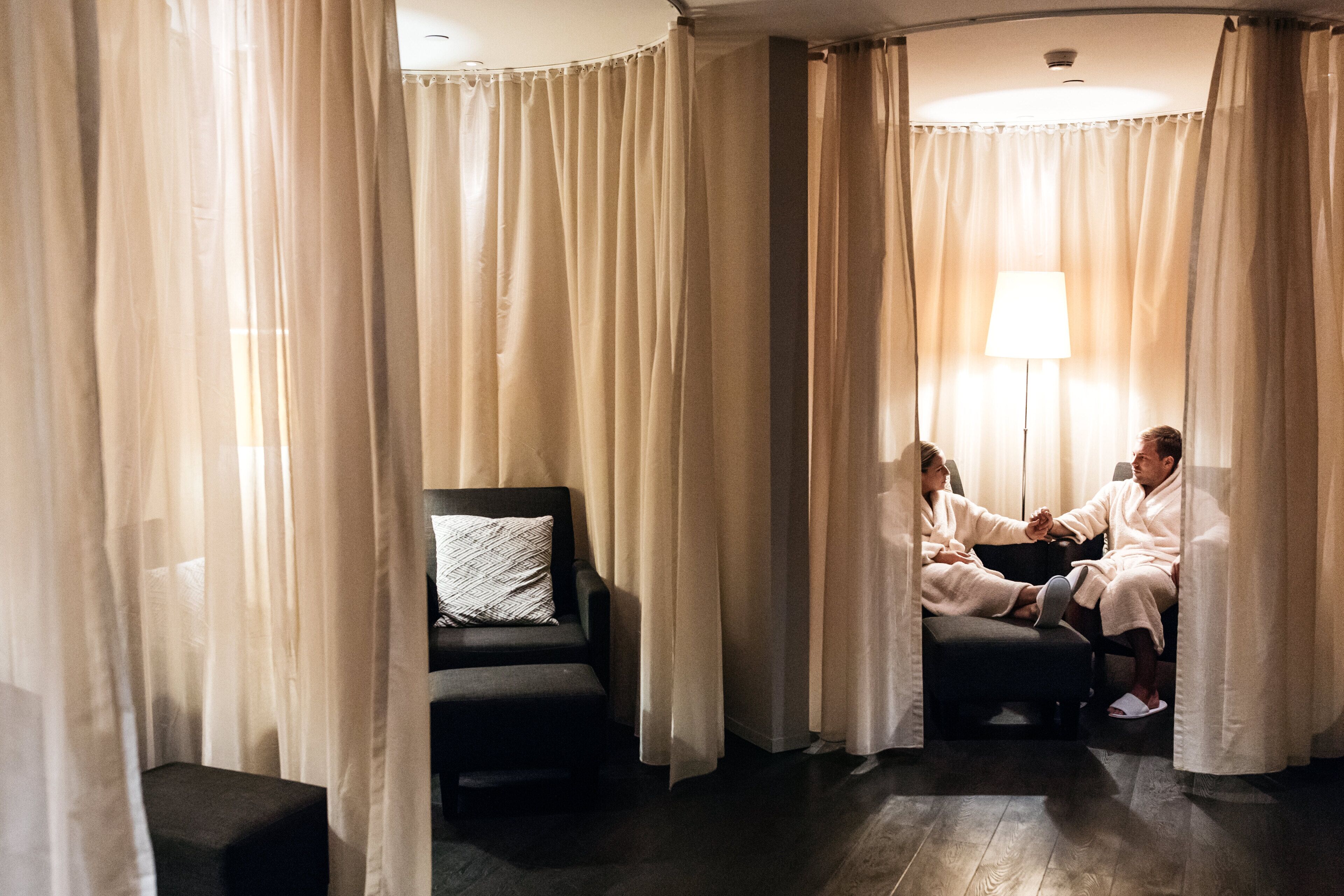 Une pièce dotée d’un parquet en bois foncé, d’un fauteuil noir avec un coussin blanc, et de deux personnes en robes assises sur un banc, face à face.
