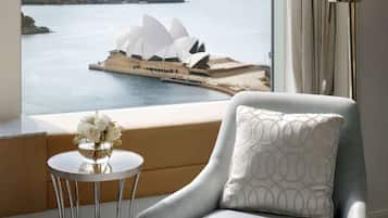 A room with a view of the Sydney Opera House, a grey armchair with a patterned pillow, a small round table with a vase of flowers, and a floor lamp.